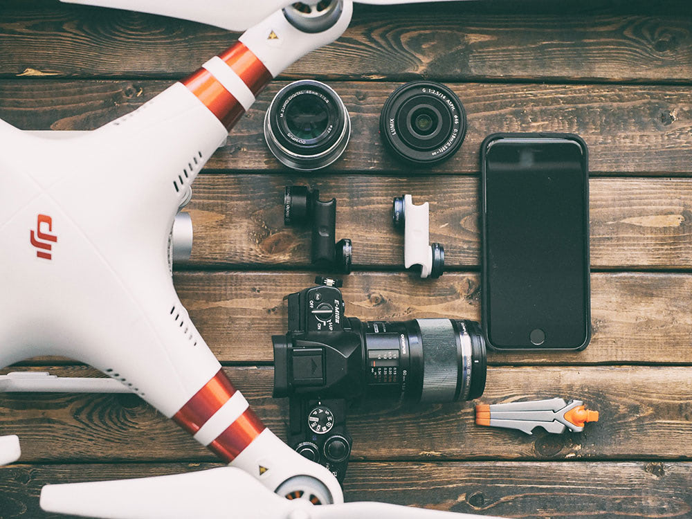 A drone, DSLR camera, lenses, smartphone, and accessories on a rustic wooden surface